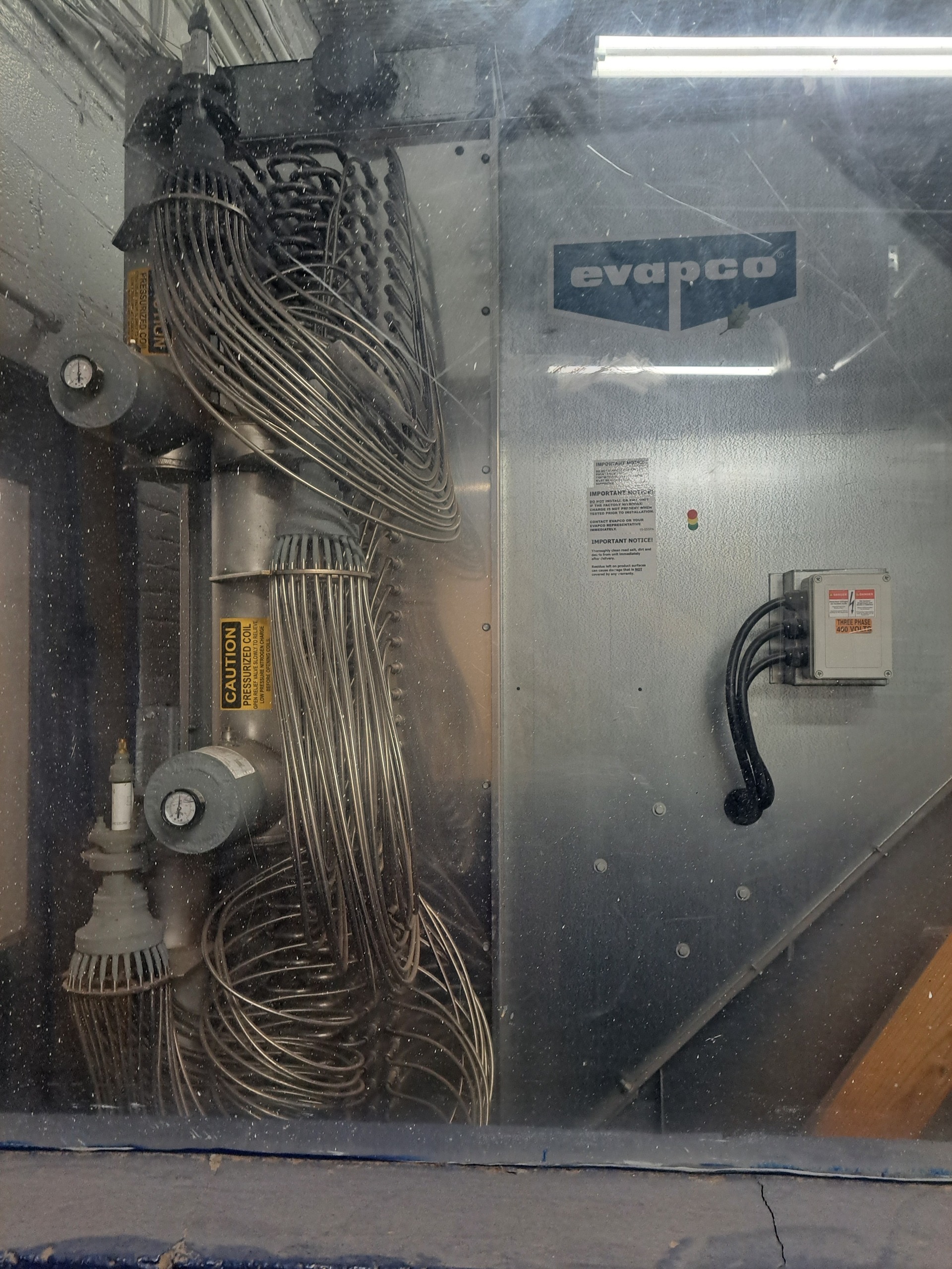 Industrial evaporative cooling unit: copper tube coil and metal housing with Evapco logo and safety labels visible outdoors/inside a facility window