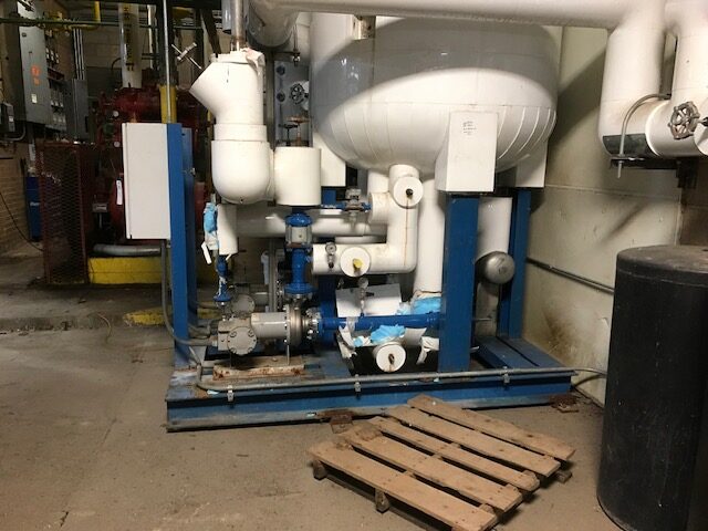 Industrial pump system with white piping and blue supports on a blue metal frame in a factory setting, near a wooden pallet.