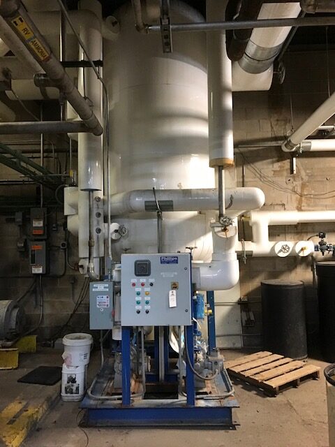 Blue skid-mounted control panel in a mechanical room, surrounded by white pipes and a tall cylindrical vessel behind it.