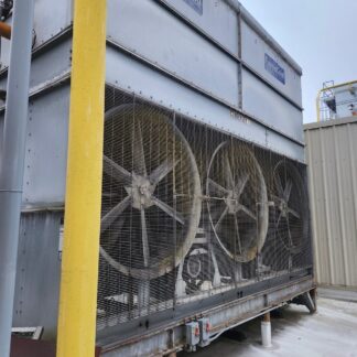 Outdoor industrial condenser unit with three large propeller fans behind a metal grille, mounted on a raised platform with piping and control boxes nearby; yellow railing post in foreground.