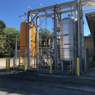 Outdoor industrial equipment with an orange vertical tank and a large silver cylinder inside a chain-link fenced area.