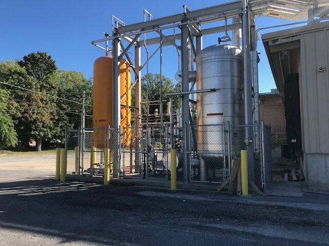 Outdoor industrial equipment with an orange vertical tank and a large silver cylinder inside a chain-link fenced area.