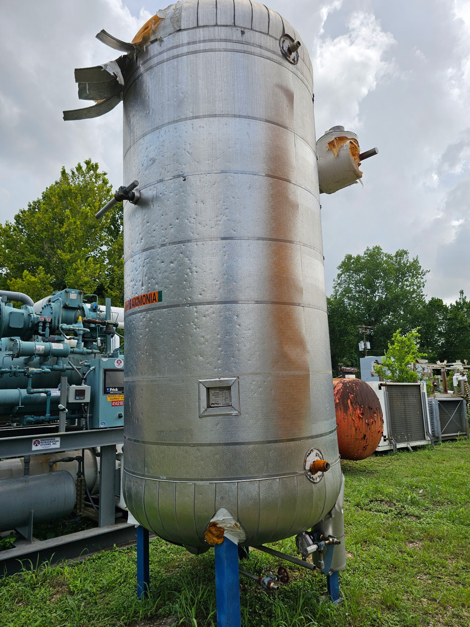 Large vertical stainless storage tank labeled ammonia, with damaged insulation and attached piping in an industrial outdoor yard.