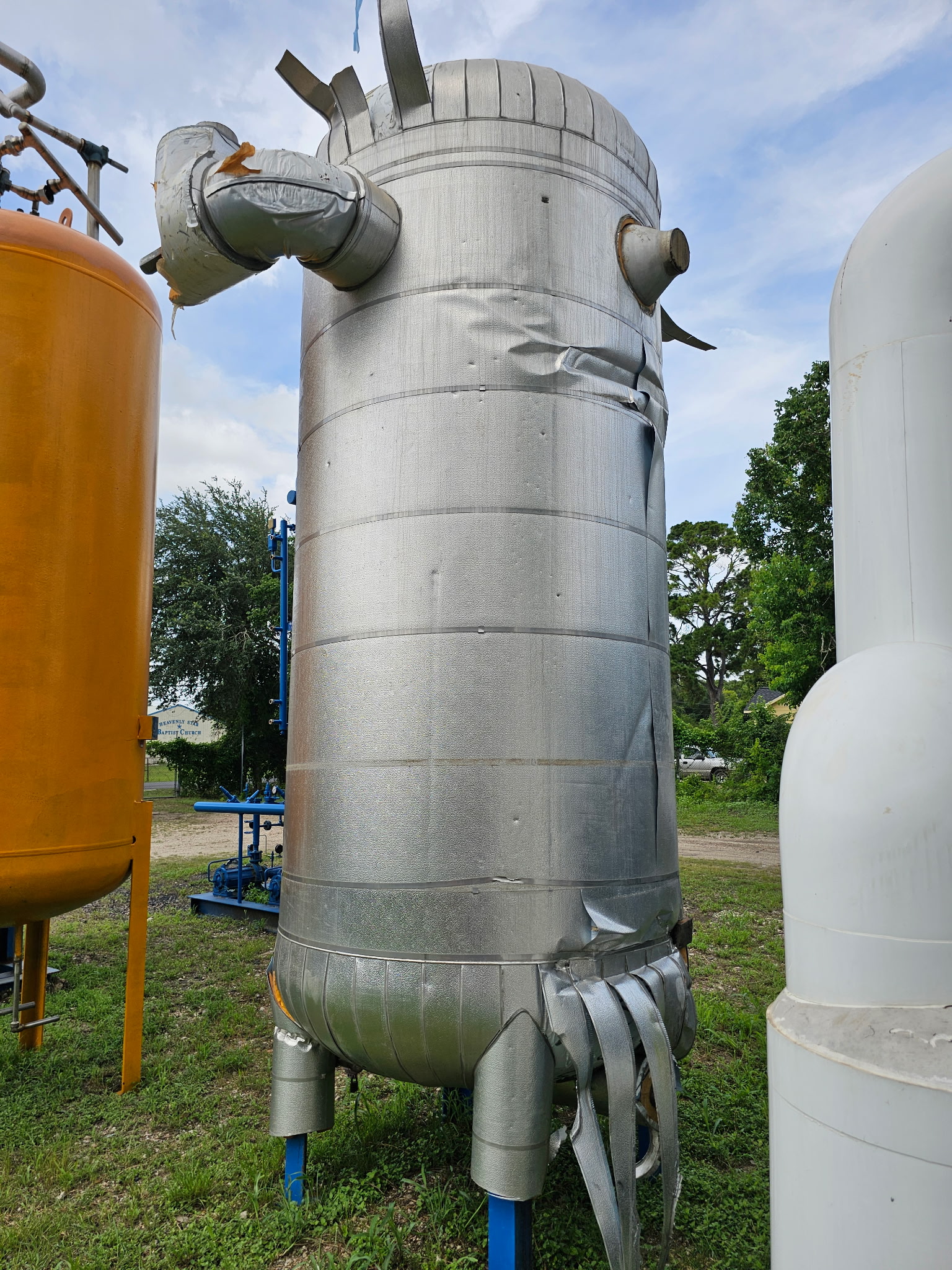 Tall silver industrial silo with wrapped ducts and protruding pipes, outdoors among other tanks.