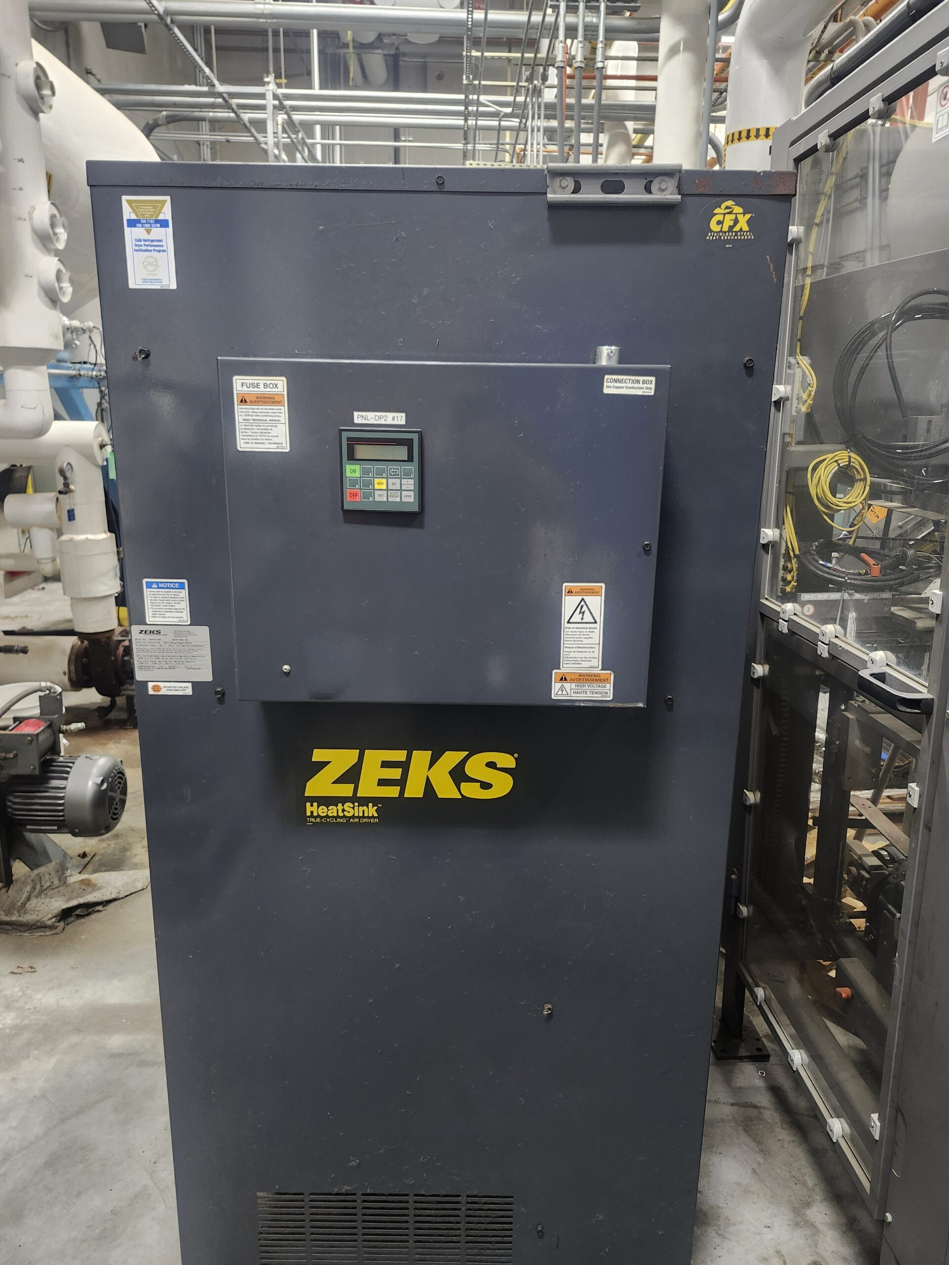 Large dark blue industrial unit labeled ZEKS HeatSink with a control panel and warning stickers, installed in a factory setting with pipes and cables overhead.