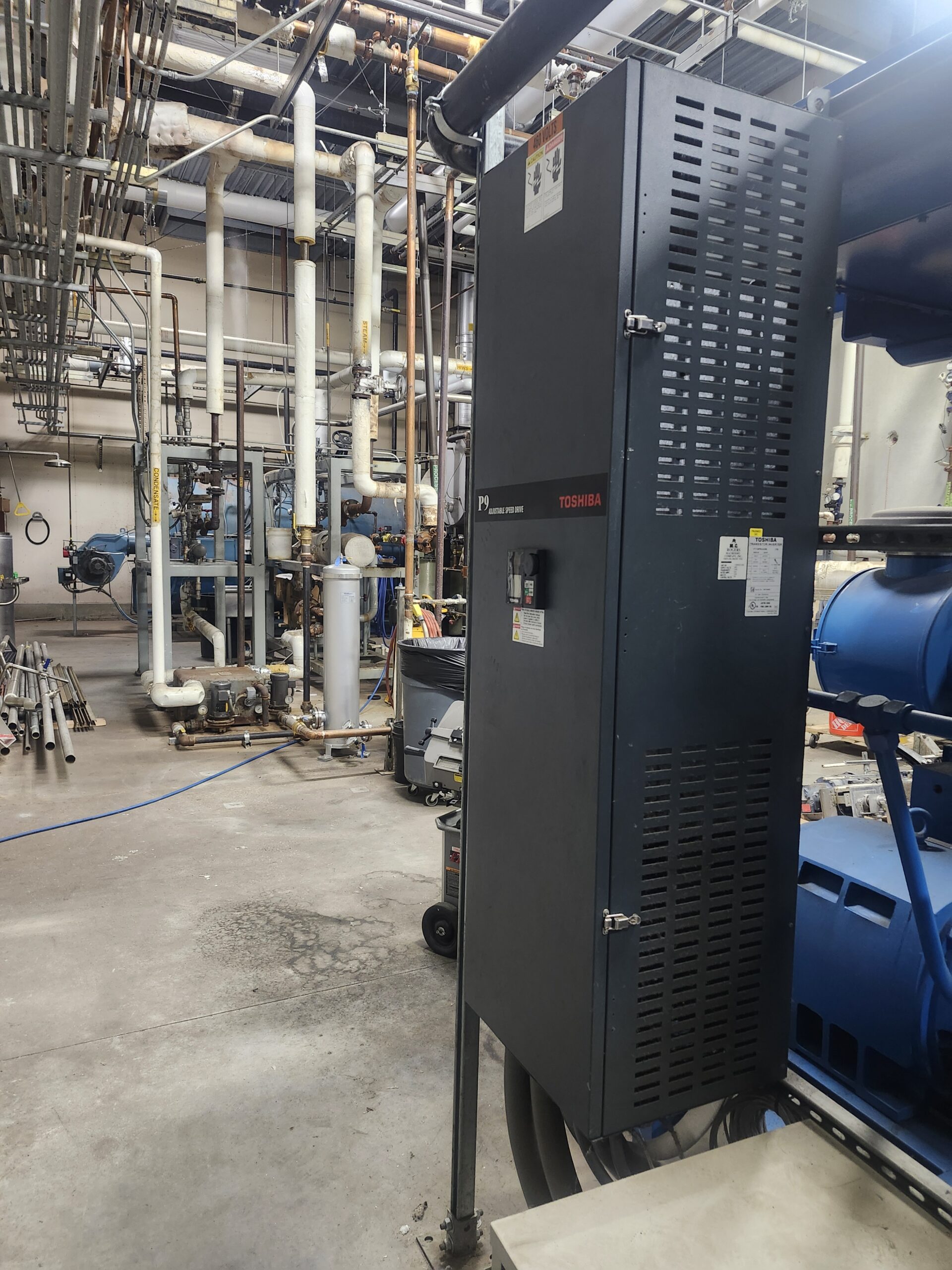 Large gray Toshiba control cabinet in a crowded utility room with numerous pipes and valves.