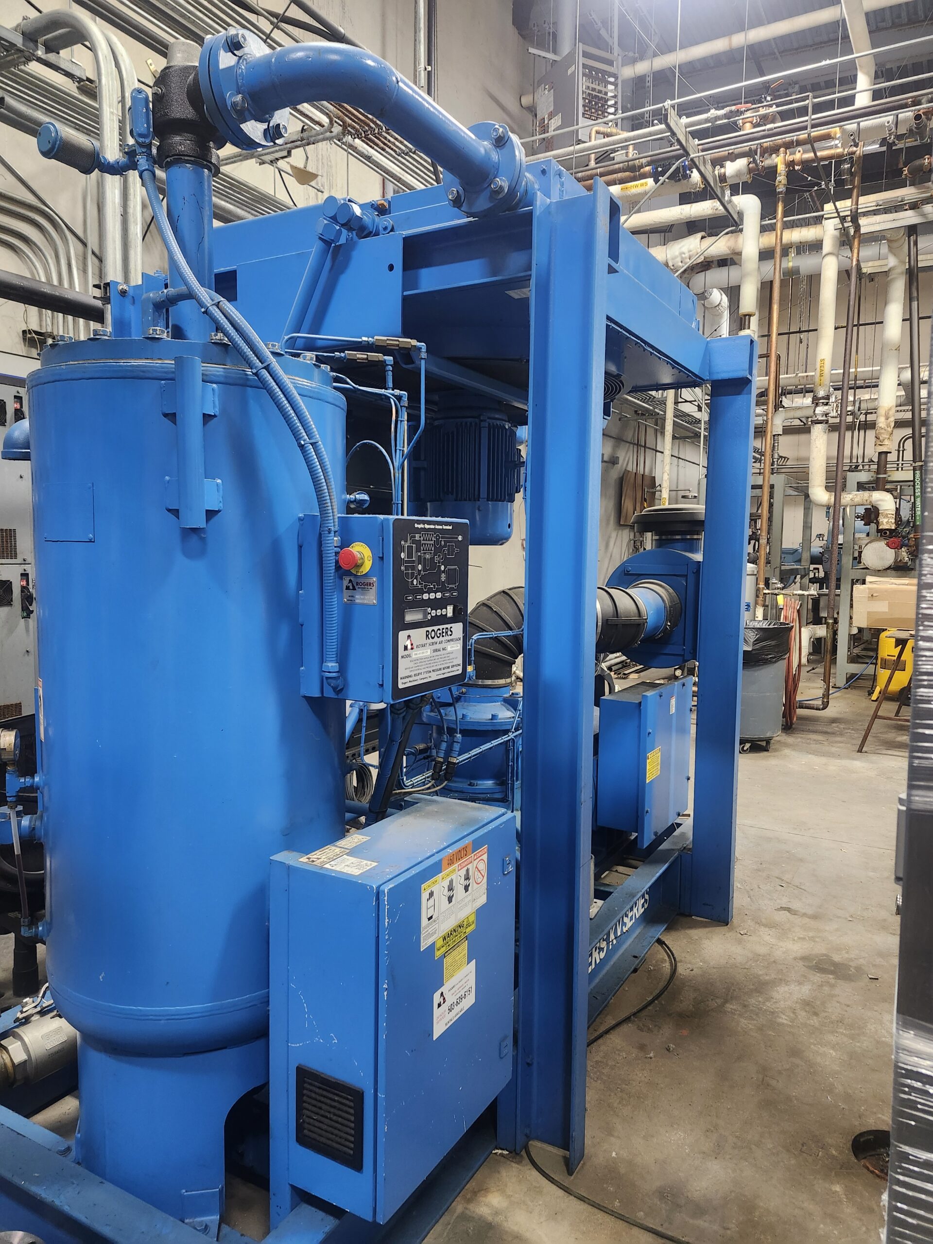 Blue industrial machinery with a large vertical tank and network of pipes in a factory utility room.