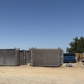 Outdoor industrial yard with stacked metal cooling units/heat exchangers, blue sky overhead and trees to the right.