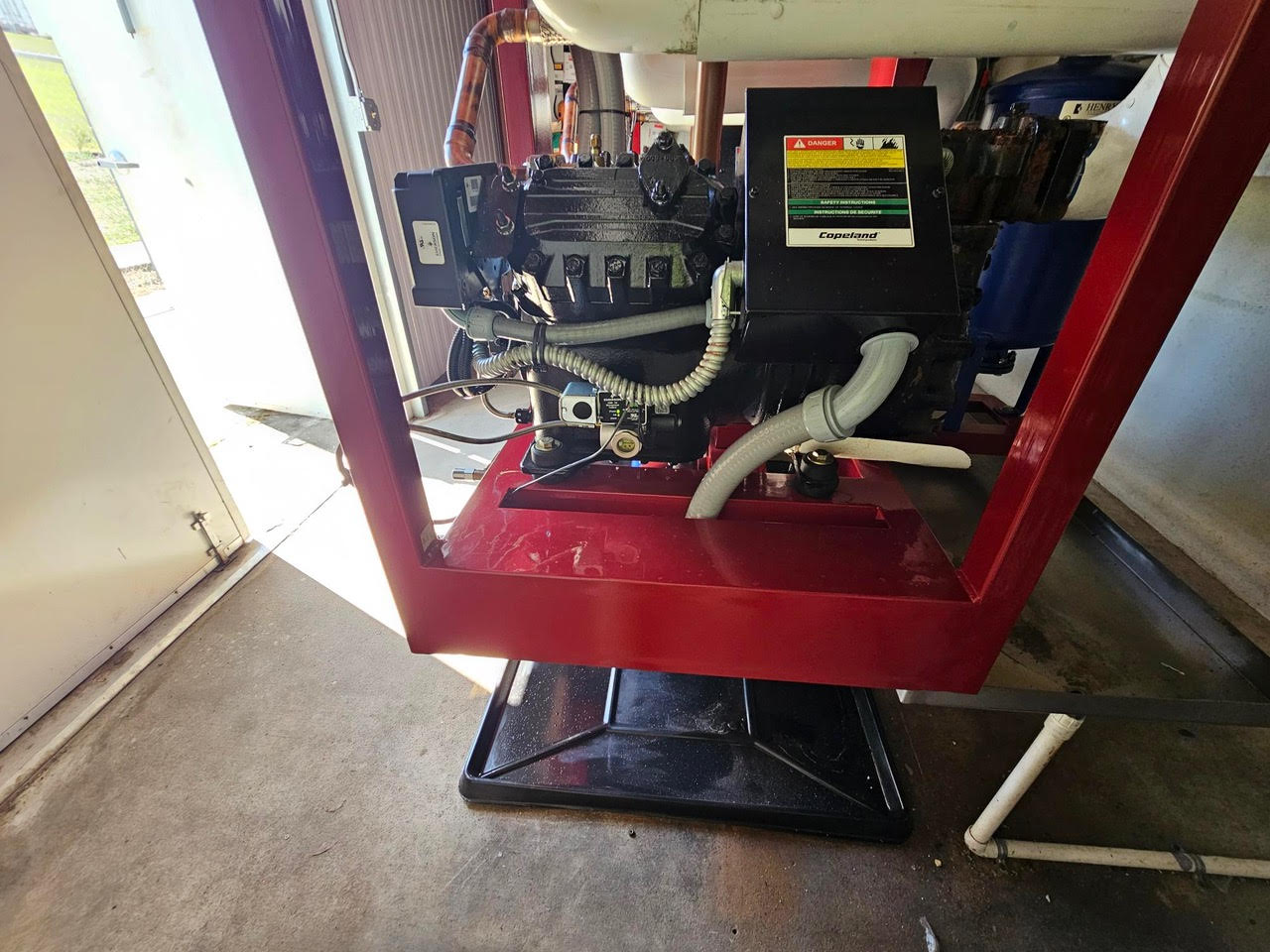 Industrial engine or generator mounted on a red frame in a workshop, with hoses, a Copeland-labeled control box, and various components visible.