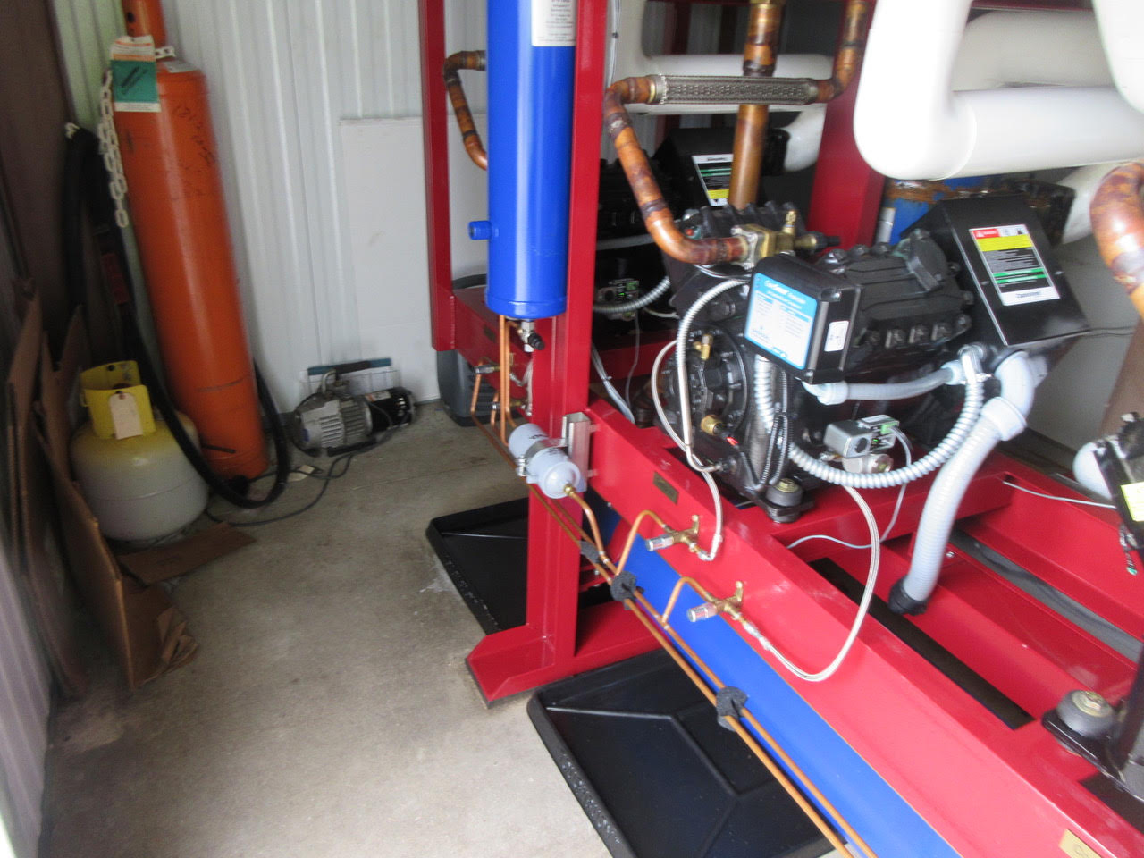 Industrial boiler system on a red frame with copper pipes and a blue vertical tank, in a workshop; propane cylinder on left.
