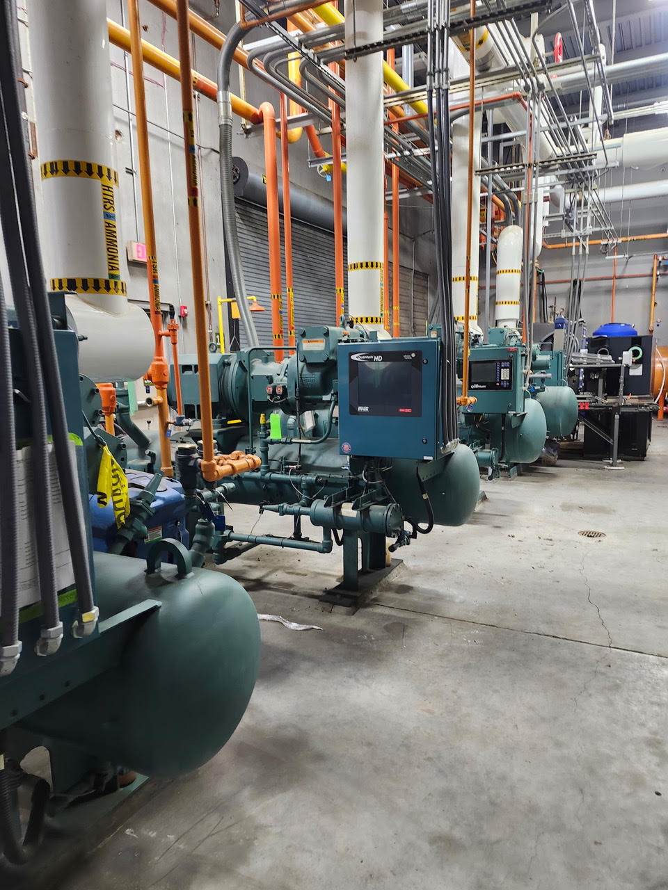Row of large green industrial generators and pumps connected by orange pipes, with yellow-black caution tape and overhead cabling in a factory setting.
