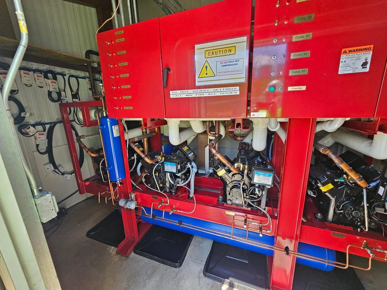 Red industrial control cabinet with warning labels, pipes, and pumps in a mechanical room.