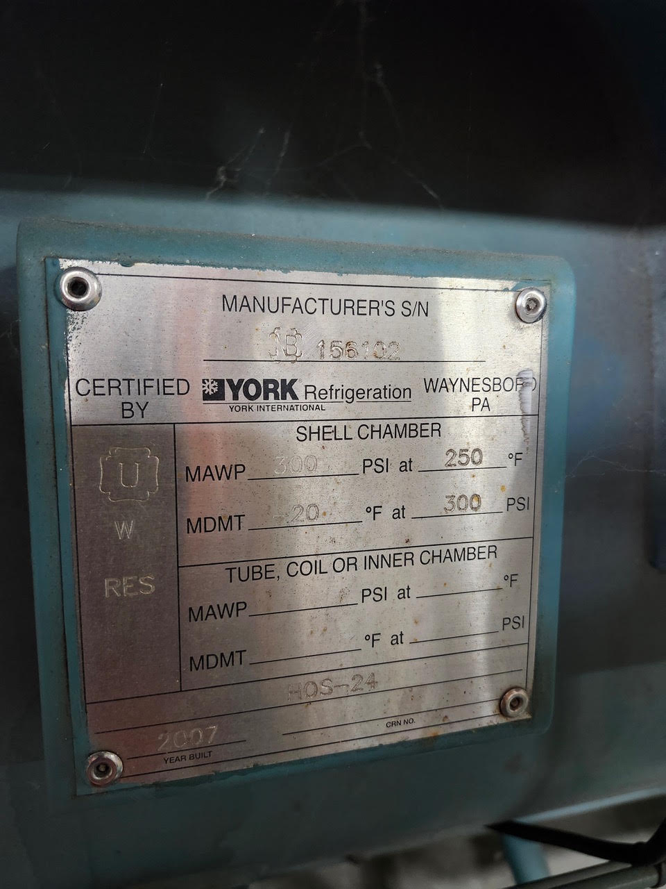 Metal nameplate on a teal machine showing manufacturing and certifying information for a York refrigeration unit, including serial number, MAWP/MDMT values, and shell chamber details (worn plate with screws).