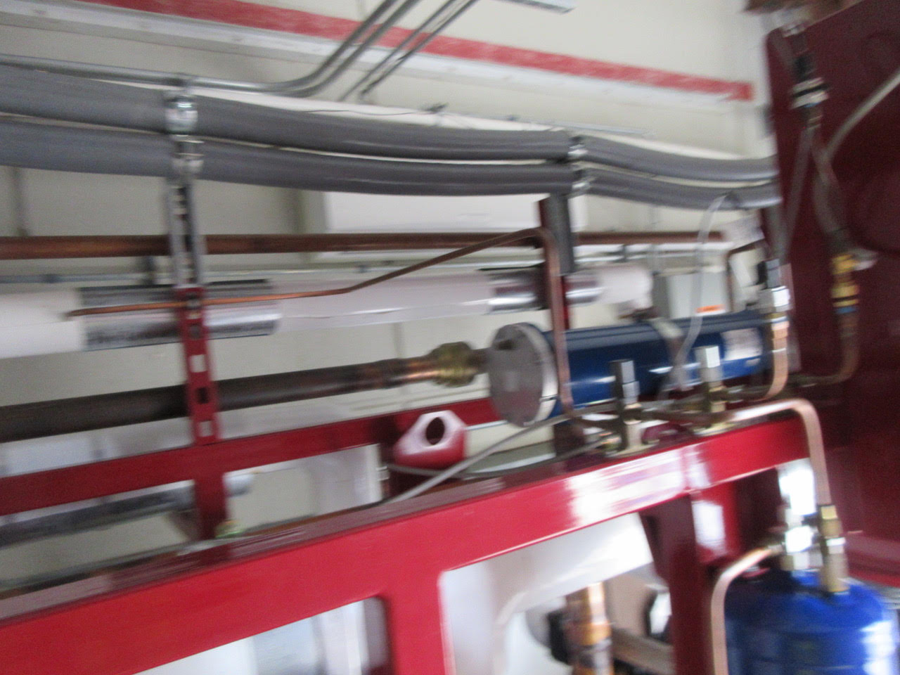 Industrial mechanical room scene showing a blue pump/motor mounted on a red frame with copper pipes and grey conduits overhead, typical of a heating or plumbing system (image is blurred).
