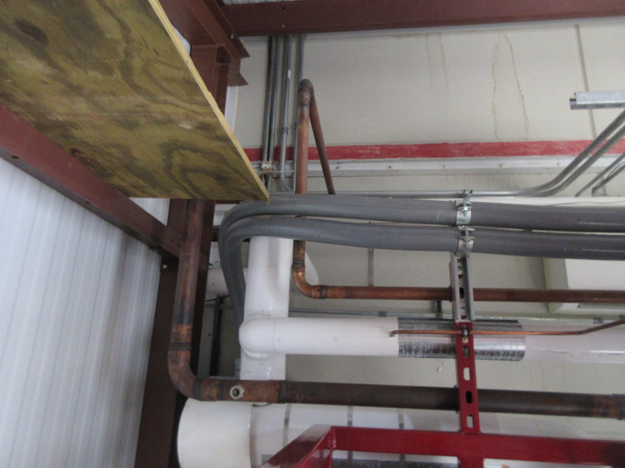 Exposed plumbing in a basement: copper and PVC pipes run along the ceiling with a wooden board and red metal support beam nearby.
