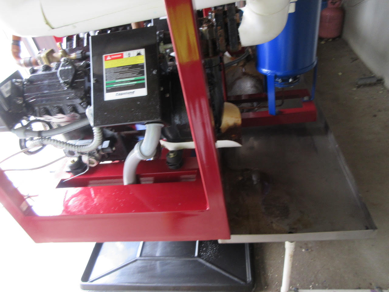 Industrial engine or boiler assembly on a red metal frame with a blue vertical tank and multiple hoses/pipes nearby.