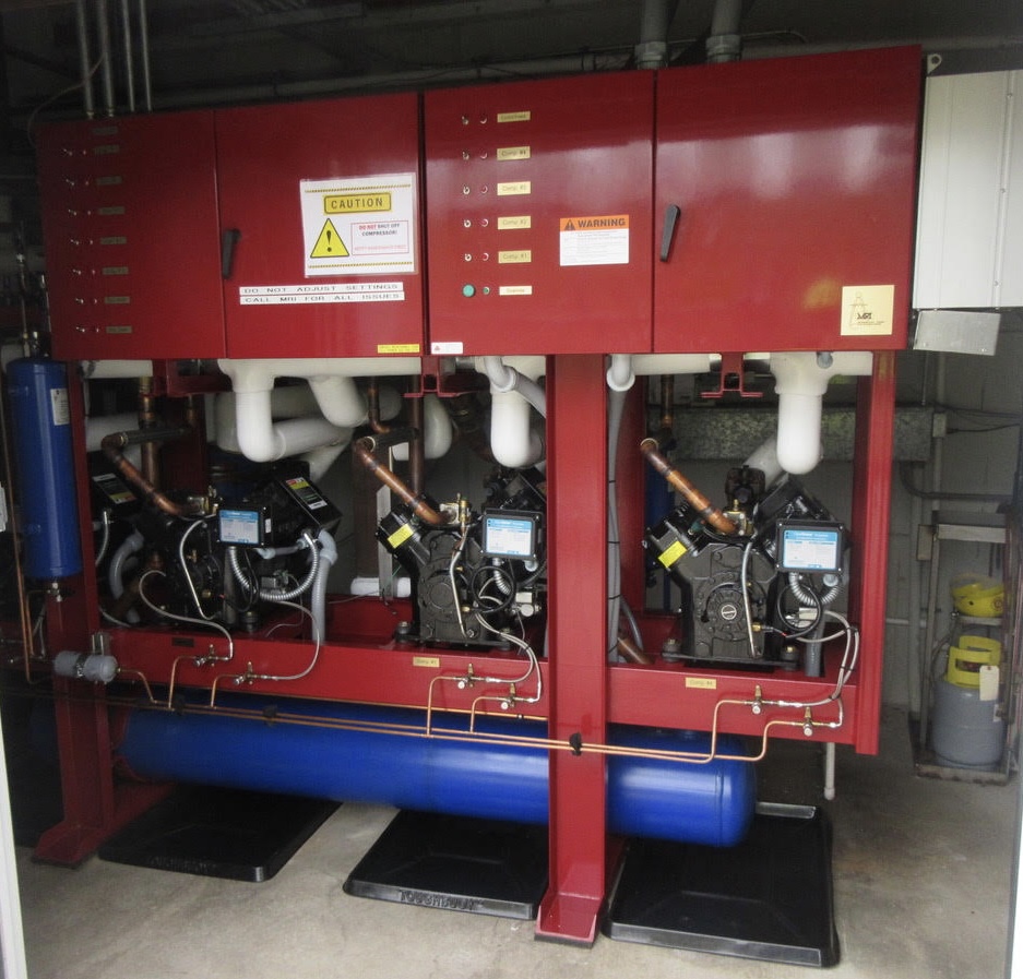 Two red industrial chiller units with white piping, pumps, and a blue reservoir beneath in a mechanical room.