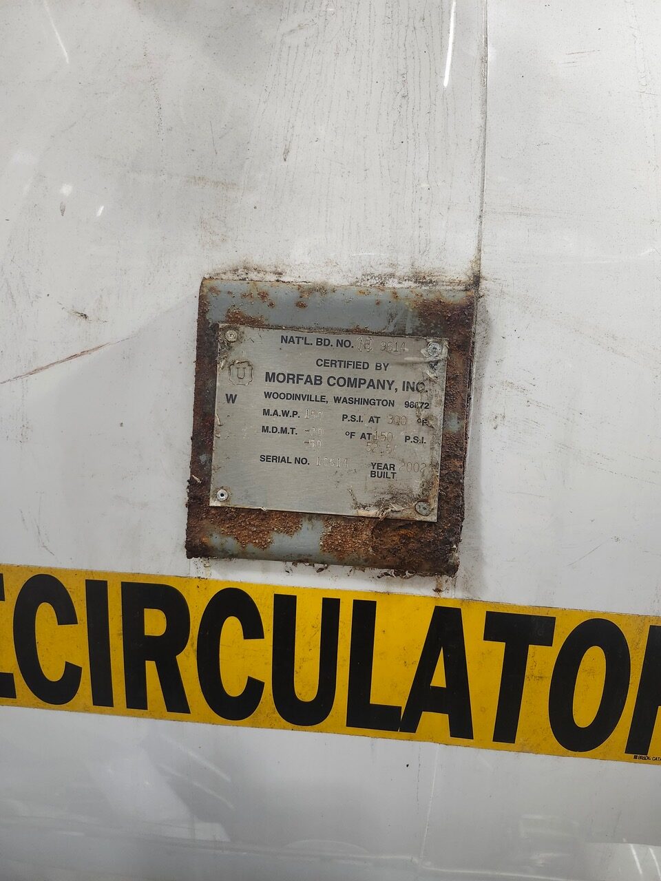 Weathered metal nameplate attached to a white surface with rust around the edges; reads 'Certified by MORFAB COMPANY, INC.' with serial number and year built, next to a yellow 'CIRCULATOR' banner.
