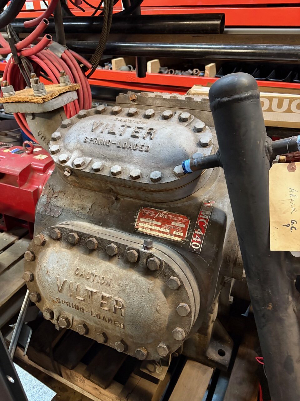 Close-up of a rugged VILTER spring-loaded valve with bolted circular covers, large flanges, and surrounding red cables in a workshop setting.
