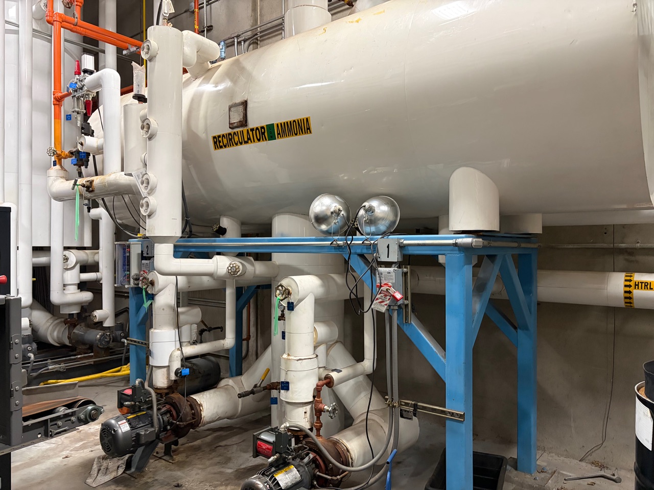 Horizontal ammonia recirculator vessel mounted on a blue steel frame, surrounded by white piping and valves in an industrial plant.