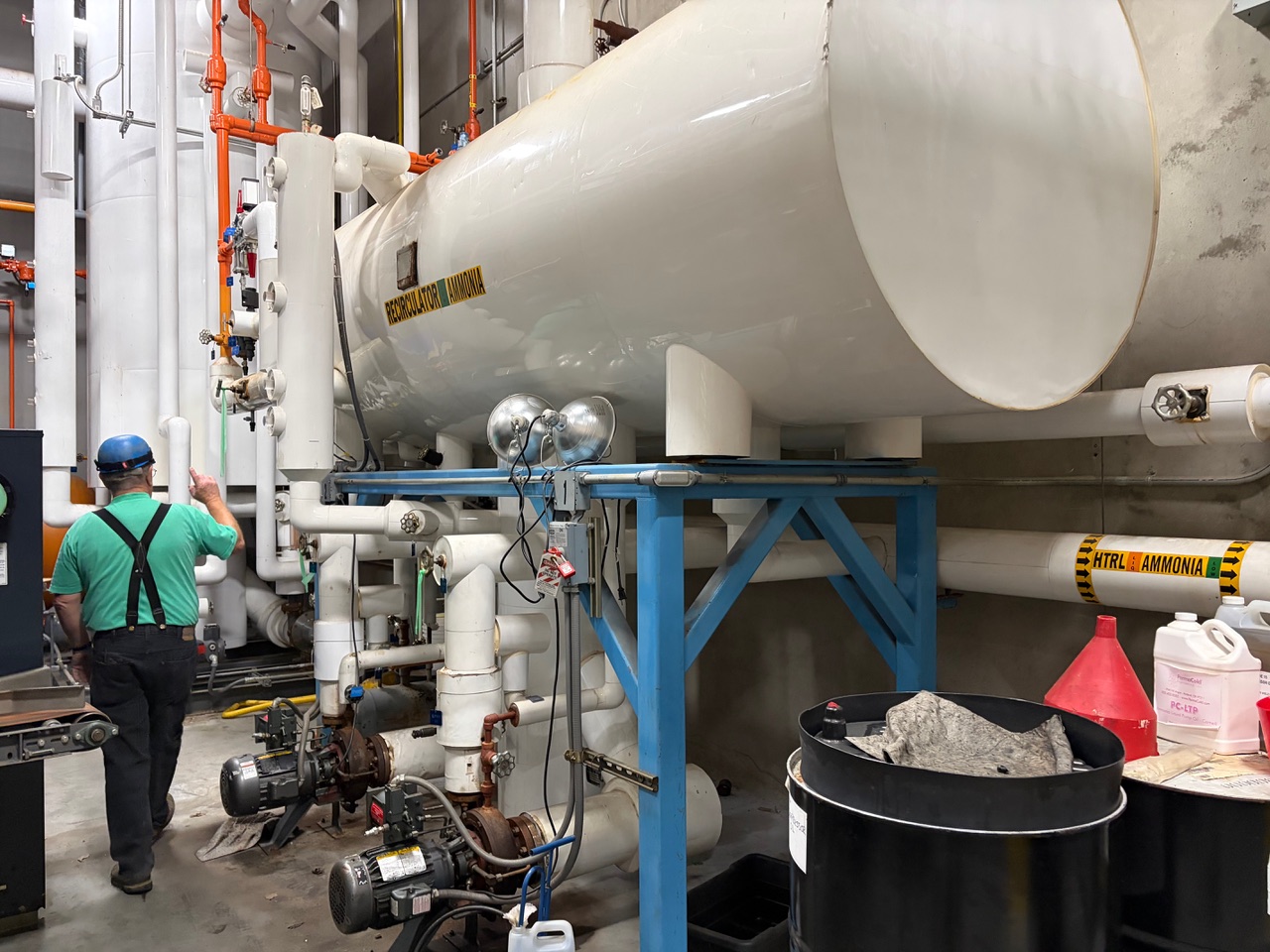 Industrial mechanical room with a large white ammonia refrigeration tank on a blue stand, worker in teal shirt and blue helmet inspecting pipes and valves.