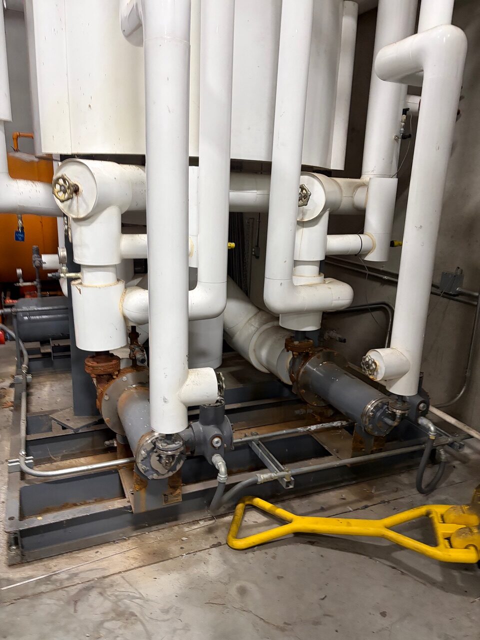 Industrial mechanical room: large white PVC pipe network with two rusted pumps and valves on a metal base. A yellow tool lies on the floor in front.
