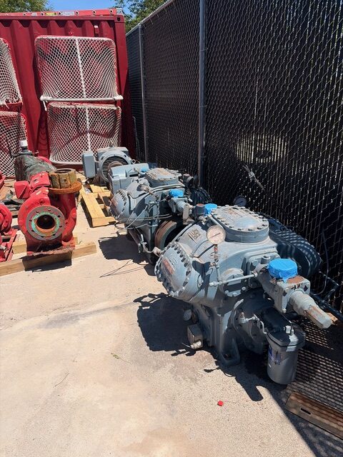 Industrial pump or compressor unit with blue covers, outdoors beside a chain-link fence and red storage containers
