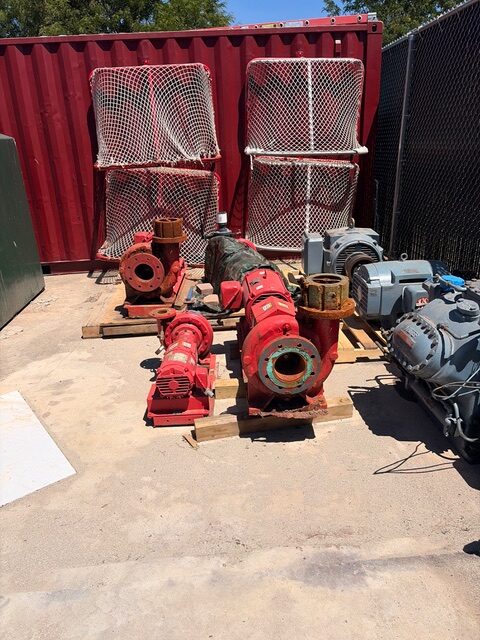 Red industrial pumps and motors arranged on wooden pallets in a shipping yard, with a red container and chain-link fence in the background.