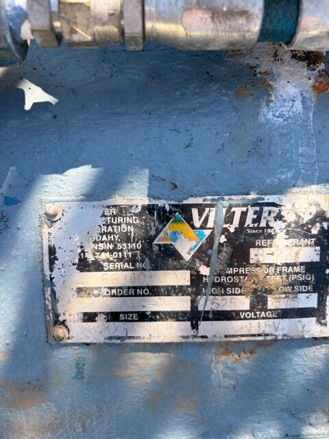 Weathered metal nameplate on blue industrial equipment showing model, serial, size, and voltage with a diamond warning symbol.