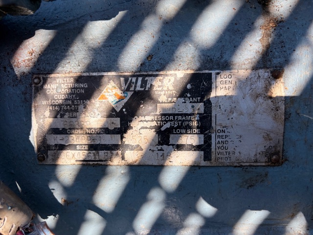 Rectangular metal nameplate on blue industrial equipment showing the faded brand 'VILTER Manufacturing Corporation' and serial numbers, partially obscured by shadows.