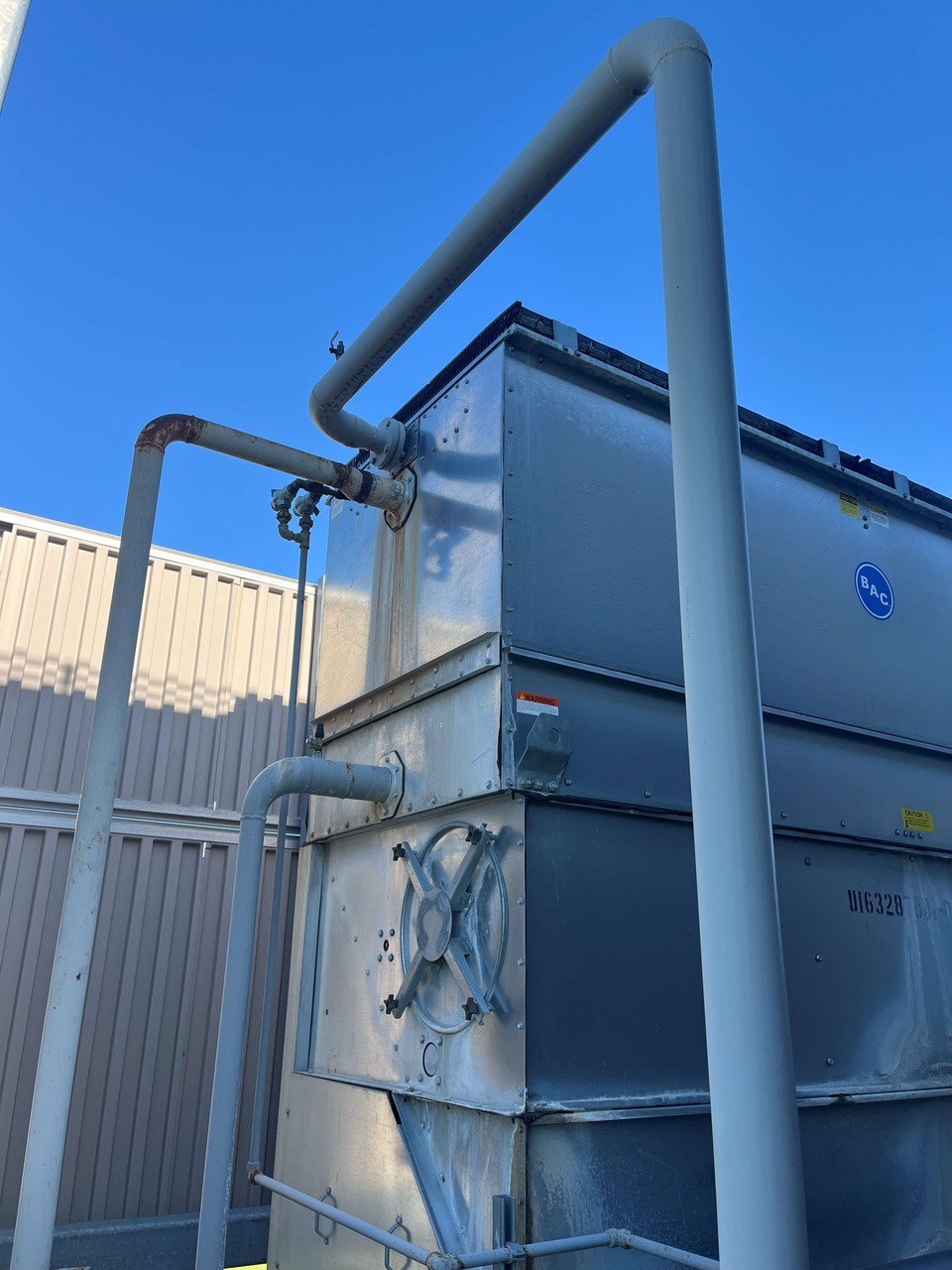 Industrial HVAC unit on a roof with silver panels, white pipes, and a large handwheel valve, BAC logo visible against a bright blue sky.