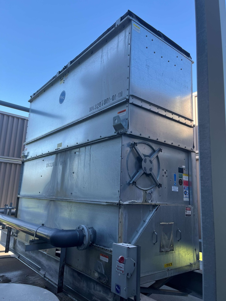 Industrial outdoor ventilation unit with a side-mounted circular fan and large ductwork, metal panels, and warning labels visible on a sunny day.