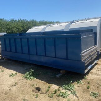 Blue metal dumpster resting on blocks in an outdoor storage yard with white equipment behind and some greenery nearby.