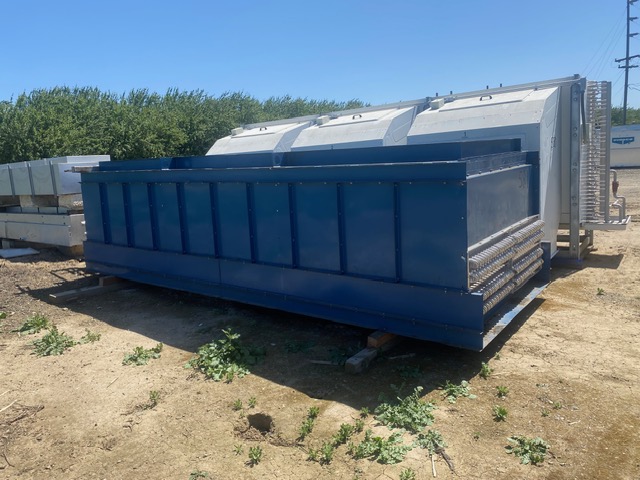 Blue metal dumpster resting on blocks in an outdoor storage yard with white equipment behind and some greenery nearby.
