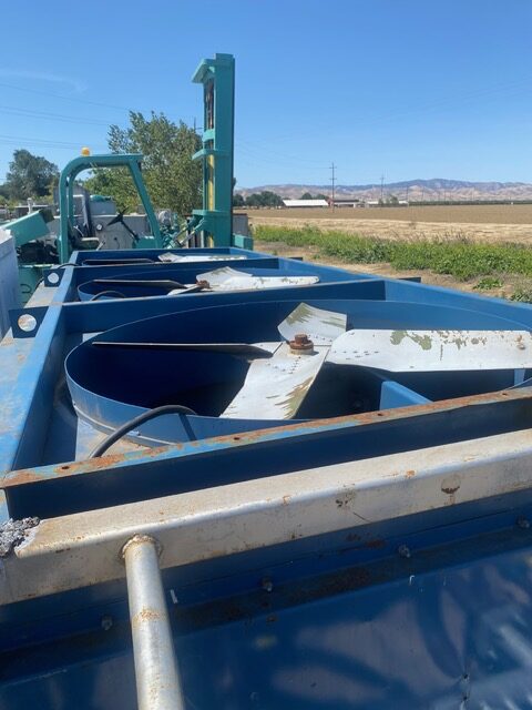 Blue agricultural machinery with large curved metal blades mounted on a trailer, parked beside an open field under a clear blue sky (farmland in distance).