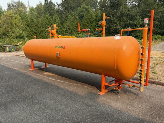 Large orange horizontal pressurized storage tank with valves and gauges on supports beside a paved lot, labeled 'RECEIVER'.