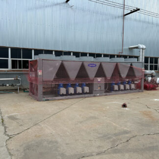 Industrial outdoor scene with a large corrugated metal building and fenced equipment enclosure; cooling unit labeled Carrier behind pink safety netting.