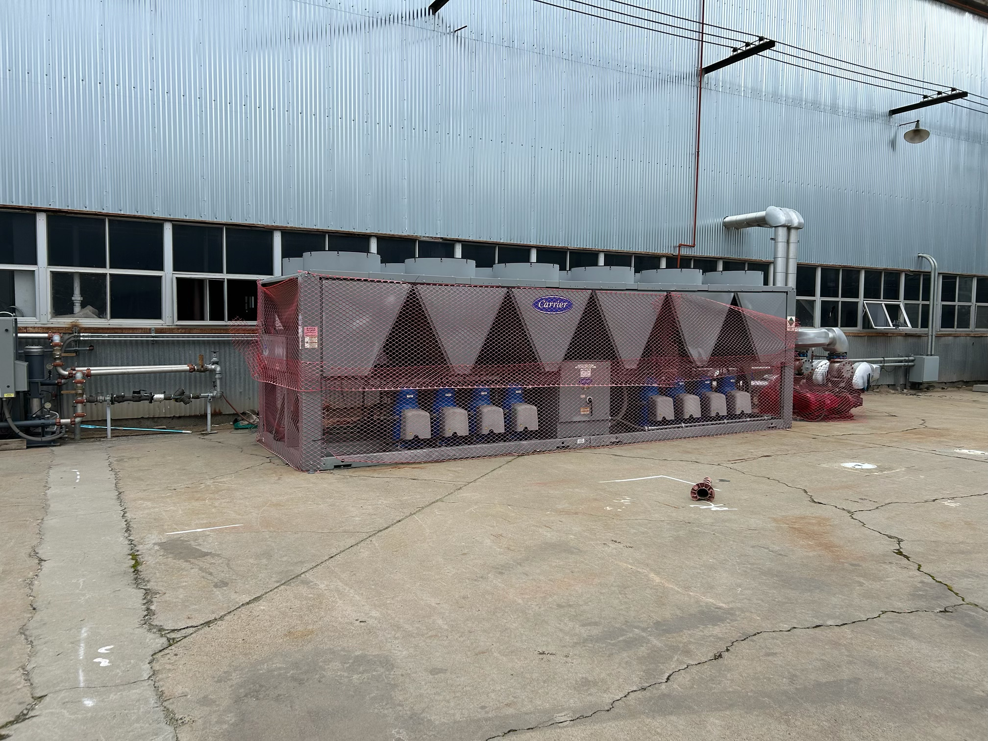 Industrial outdoor scene with a large corrugated metal building and fenced equipment enclosure; cooling unit labeled Carrier behind pink safety netting.