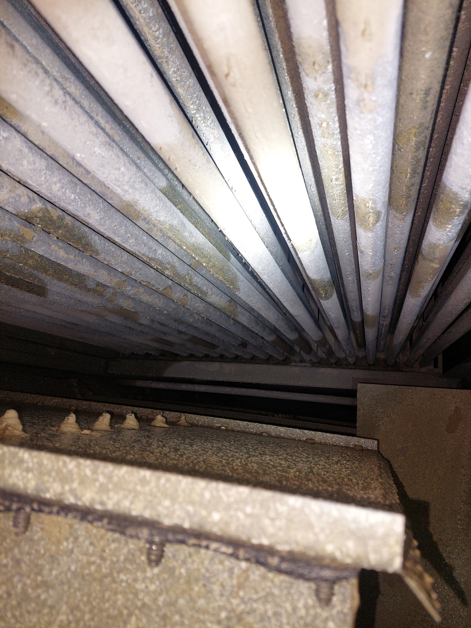 Interior view of a dusty, rust-speckled metal duct or vent with parallel bars/ribs forming a circular pattern.