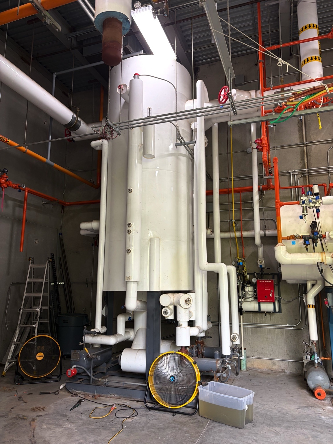Tall white cylindrical industrial vessel with numerous white pipes, red/orange piping, and valves in a mechanical room
