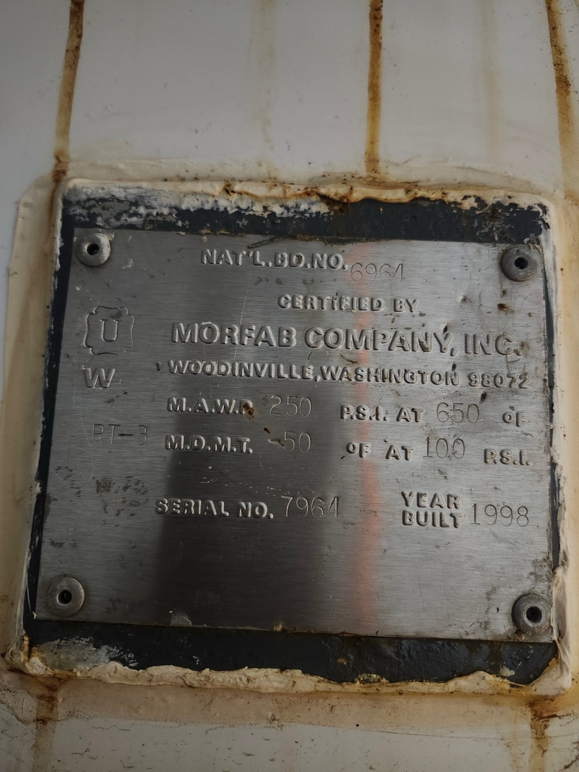 Rusted metal nameplate attached to a surface; reads MORFAB COMPANY INC, Woodinville WA, serial no. 7964, year built 1998, with worn edges and screws visible.