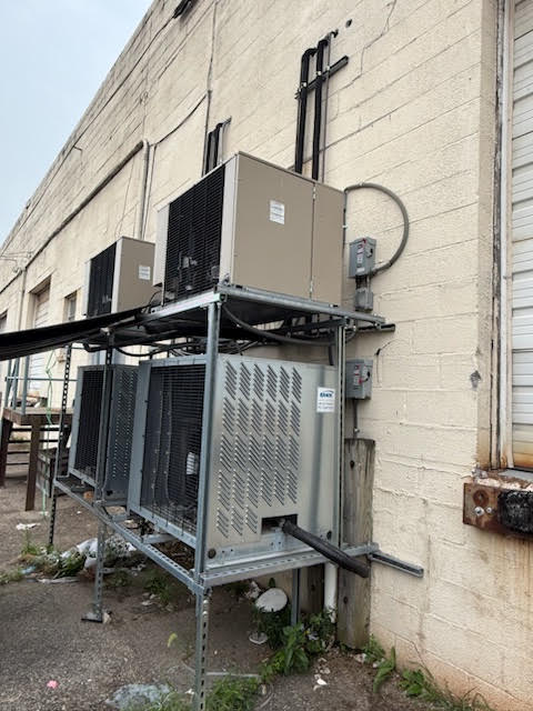 Row of outdoor commercial HVAC units on a metal platform attached to a beige brick wall, with electrical conduits and ductwork.