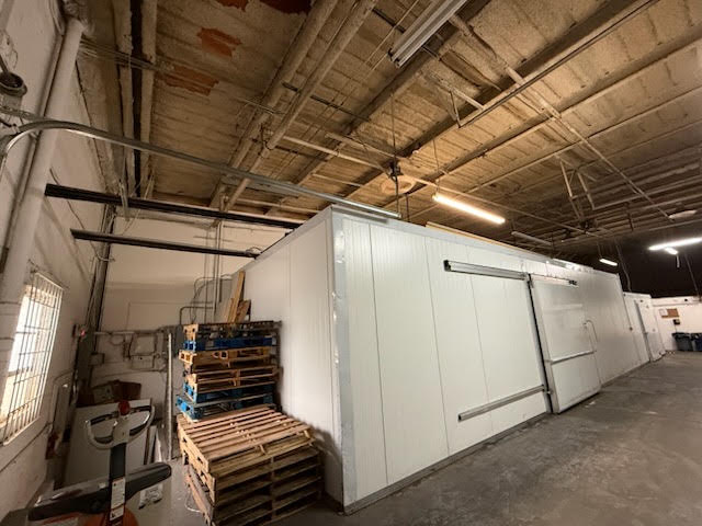 Industrial warehouse interior with white insulated storage modules, pallets, and exposed ceiling pipes.