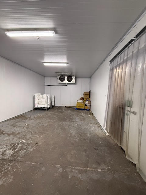 Industrial cold storage room with white walls, concrete floor, overhead lights, curtain on the right, and pallets of boxes near the far end.