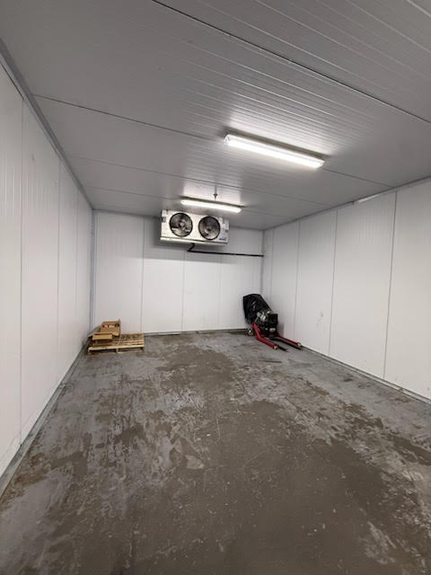 Empty industrial room with white insulated walls and bare concrete floor; overhead lights, a wooden pallet on the left, and red equipment on the right near a wall-mounted cooling unit.