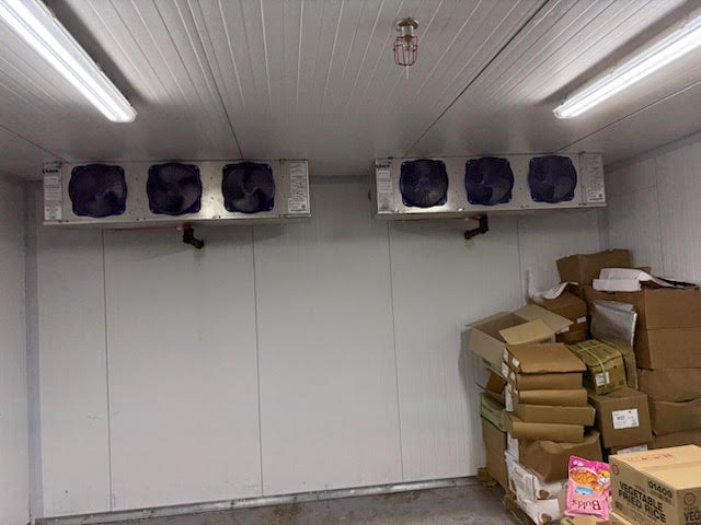 Cold storage room with white insulated walls, two ceiling-mounted cooling units, and a stack of cardboard boxes on the right under fluorescent lights.