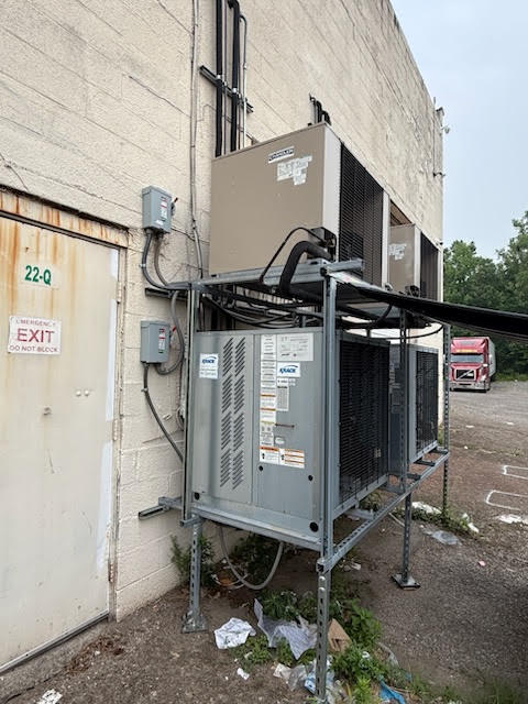Outdoor HVAC condenser unit on a metal frame beside a beige building, with piping and electrical boxes; a truck visible in the background.